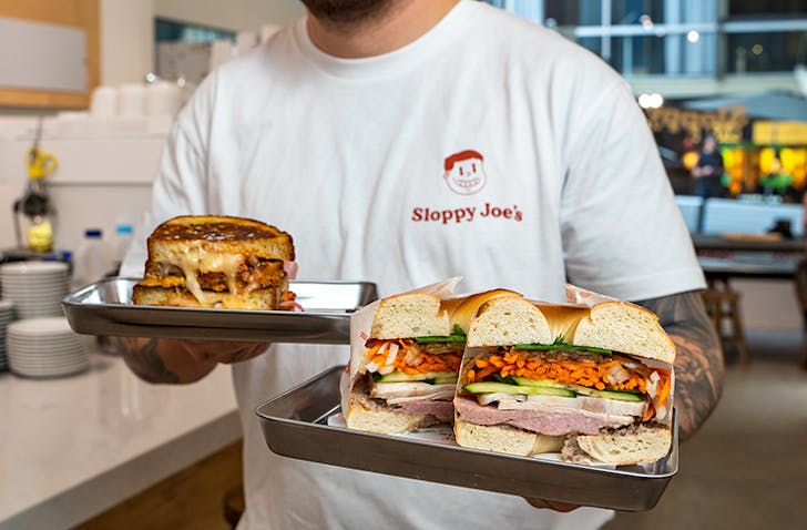 A man holding a large bagel and one of Melbourne's best sandwiches in a white tshirt.